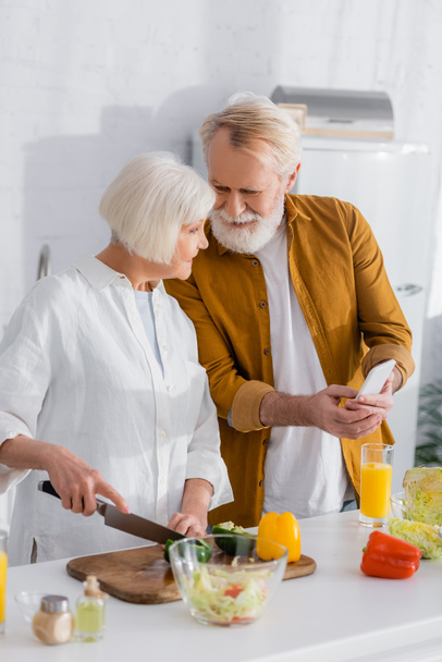 Senior mężczyzna gospodarstwa smartfon w pobliżu uśmiechnięta żona gotowanie na zamazanym pierwszym planie w kuchni  - Zdjęcie, obraz