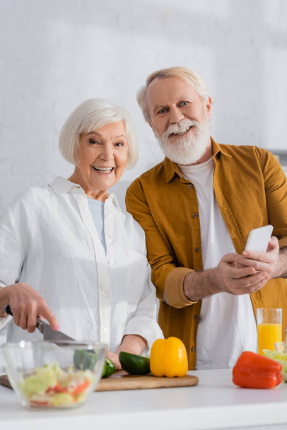 Улыбающаяся пожилая пара со смартфоном, смотрящая в камеру во время приготовления пищи на размытом переднем плане  - Фото, изображение