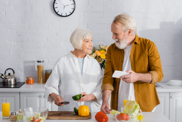 Senior man met smartphone in de buurt van glimlachende vrouw met mes en paprika in de keuken  - Foto, afbeelding