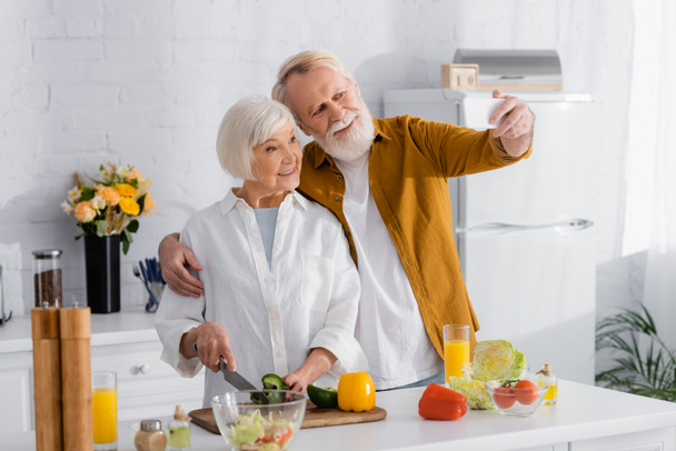Senior man taking selfie on smartphone with smiling wife cooking in kitchen  - Foto, imagen