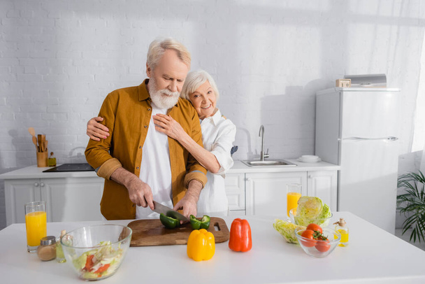 Senior woman hugging husband cooking near orange juice in kitchen  - Fotó, kép