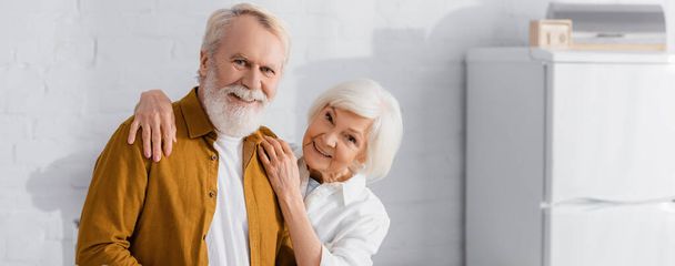 Χαμογελαστή ηλικιωμένη γυναίκα αγκαλιάζει τον άντρα της στην κουζίνα, πανό  - Φωτογραφία, εικόνα