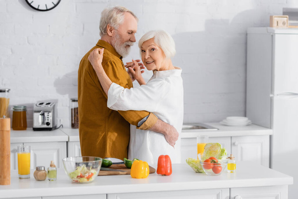 Ηλικιωμένη γυναίκα χαμογελά στην κάμερα ενώ χορεύει με τον σύζυγο κοντά σε λαχανικά στην κουζίνα  - Φωτογραφία, εικόνα