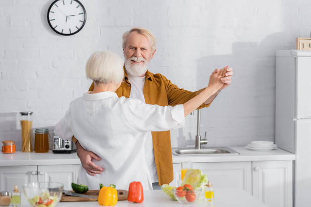 笑顔シニア男ダンスととともに妻近づい野菜上のぼやけたフォアグラウンドでキッチン  - 写真・画像