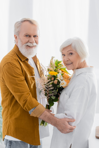 Ηλικιωμένο ζευγάρι με λουλούδια χαμογελά στην κάμερα στην κουζίνα  - Φωτογραφία, εικόνα