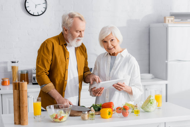 Ηλικιωμένη γυναίκα με ψηφιακό tablet στέκεται κοντά στο σύζυγο μαγείρεμα στην κουζίνα  - Φωτογραφία, εικόνα