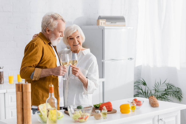 Smiling senior couple holding glasses of wine embracing near vegetables on blurred foreground  - Photo, Image