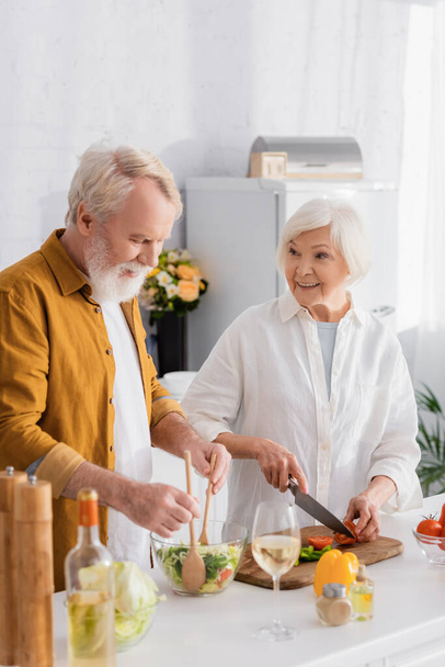 Homme âgé mélangeant salade près femme souriante et vin au premier plan flou  - Photo, image