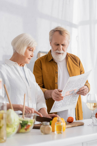 Ouderen paar lezen nieuws tijdens het koken op wazig voorgrond in de keuken  - Foto, afbeelding