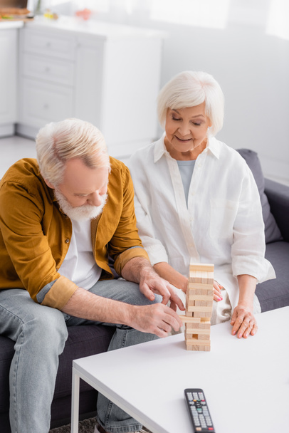 Senior άνθρωπος παίζει μπλοκ παιχνίδι ξύλο με τη σύζυγο κοντά απομακρυσμένο ελεγκτή σε θολή πρώτο πλάνο  - Φωτογραφία, εικόνα