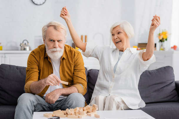 Aufgeregte Seniorin schaut Ehemann an, der einen Teil der Holzblöcke hält Spiel  - Foto, Bild