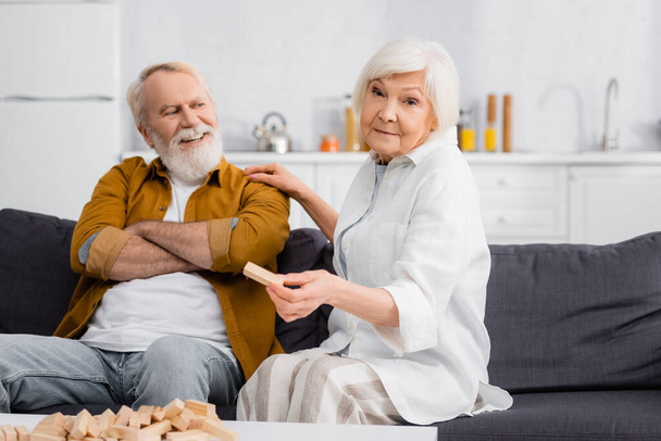 Seniorin hält Teil von Holzklötzen neben fröhlichem Ehemann auf Couch  - Foto, Bild