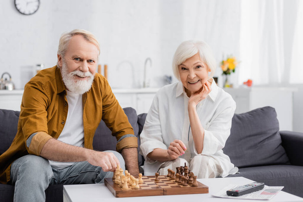 Smiling couple looking at camera near chess on coffee table  - Photo, Image