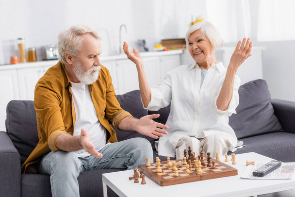 Hombre mayor señalando con las manos a los jabalíes de ajedrez cerca de la esposa sonriente en el sofá  - Foto, imagen