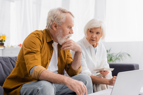 Pensativo hombre mayor sentado cerca de la esposa con calculadora y portátil en primer plano borrosa  - Foto, Imagen