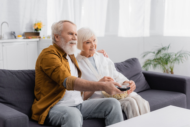 Lächelndes älteres Paar vor dem Fernseher mit Popcorn auf der Couch  - Foto, Bild