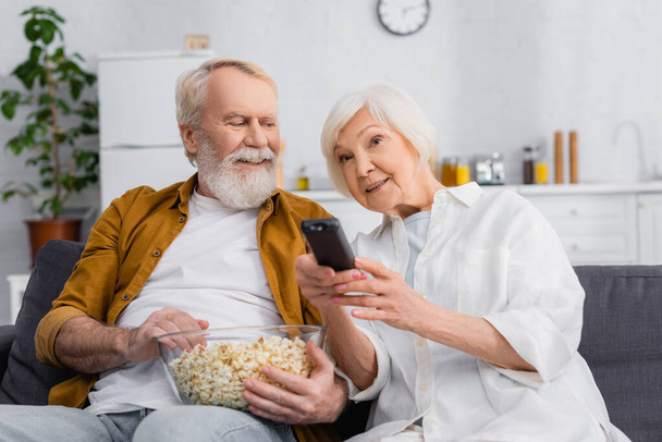 Seniorin klickt neben Ehemann mit Schüssel Popcorn auf Couch  - Foto, Bild
