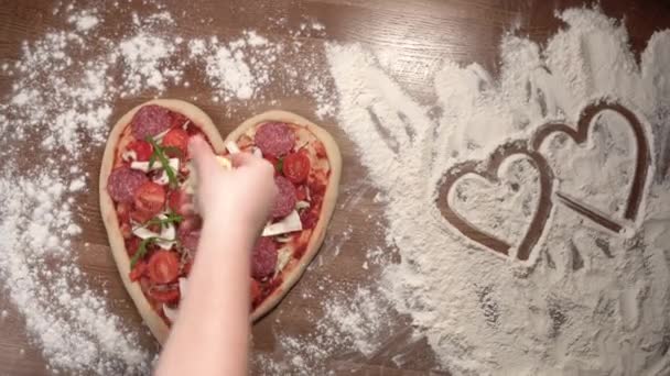 una pizza grande en forma de corazón yace sobre la mesa, una mano de mujer está poniendo queso encima. Forma de corazón, concepto de cena para el día de San Valentín, aniversario, con amor - Metraje, vídeo