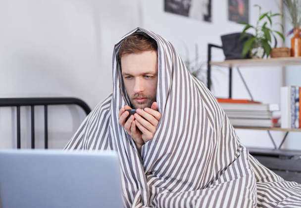 Feeling sick, healthcare, technology and remote work concept - sick caucasian bearded man wrapped with blanket having video call on laptop computer at home office - Foto, Imagen