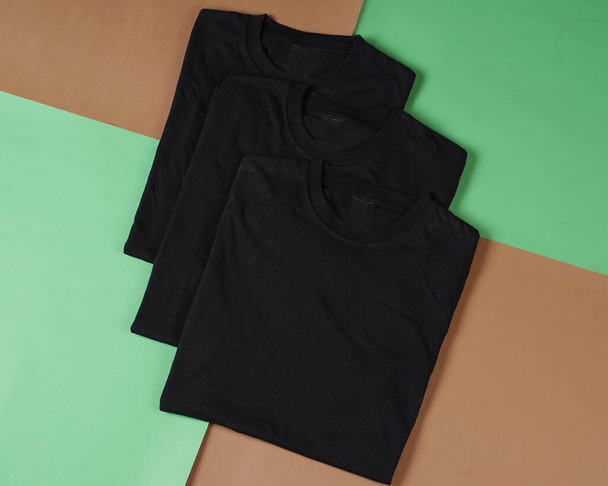 黒い折り畳まれた服の山。折り畳まれた黒いTシャツの上のビューは、色の背景、コピースペース、フラットレイアウトで隔離された。空のTシャツテンプレートは、一般的にモックアップやテンプレートデザインに使用されます. - 写真・画像