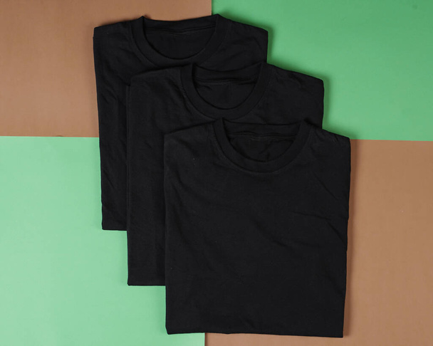 Stapel schwarzer Klamotten. Draufsicht auf gefaltetes schwarzes T-Shirt isoliert mit farbigem Hintergrund, Kopierraum, flache Lage. Leere T-Shirt-Vorlagen werden häufig für Attrappen und Vorlagendesigns verwendet. - Foto, Bild