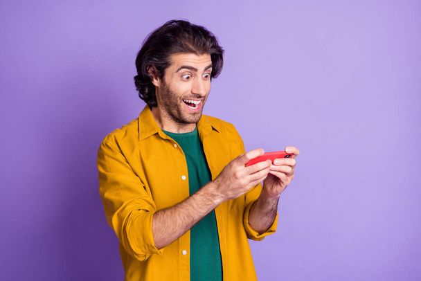 Photo of young man blogger excited happy hold smartphone chat read like post wear yellow shirt isolated over purple color background - Photo, image