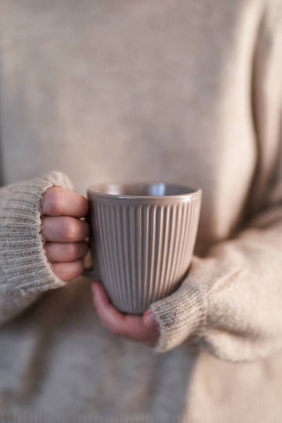 Manos femeninas en jersey beige sosteniendo taza de café caliente, chocolate caliente o té. Concepto de confort invernal, por la mañana, sensación cálida - Foto, imagen