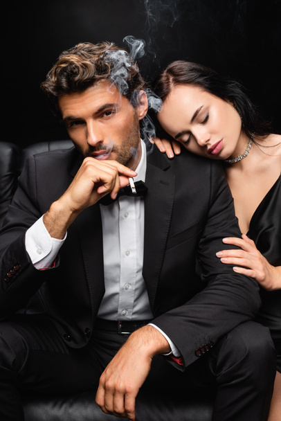 sensual woman with closed eyes hugging shoulder of elegant man smoking and looking at camera isolated on black - Photo, image