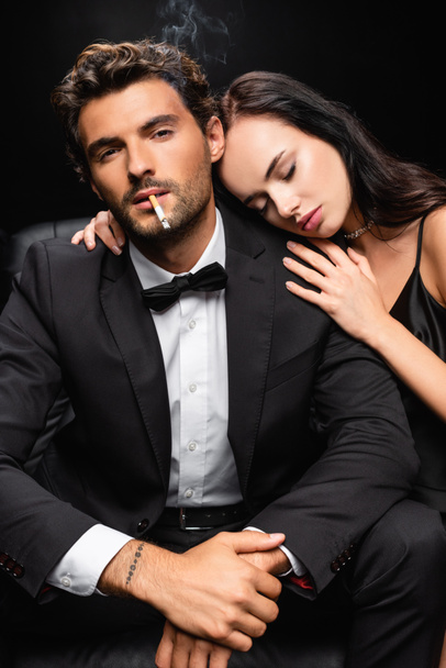 seductora morena apoyada en el hombro del hombre fumando y mirando a la cámara aislada en negro - Foto, imagen