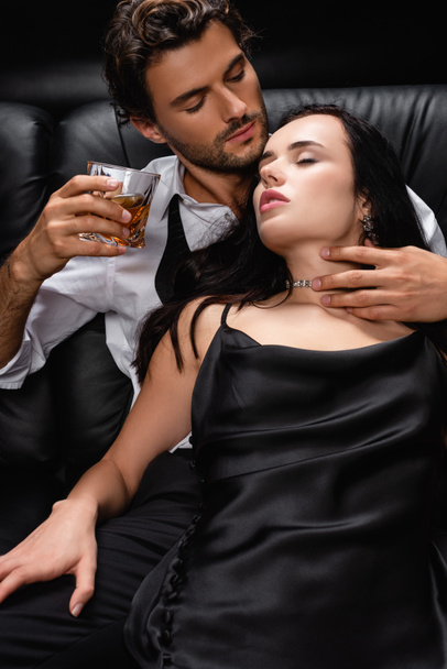 young man holding glass of whiskey and embracing neck of seductive woman on black - Фото, изображение