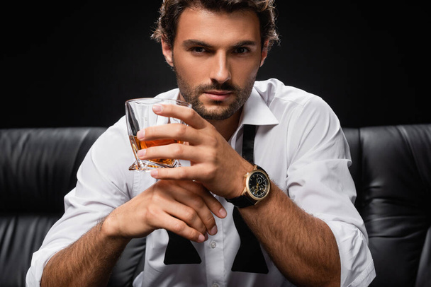 man with glass of whiskey, in white shirt and untied tie, looking at camera isolated on black - Foto, Imagen