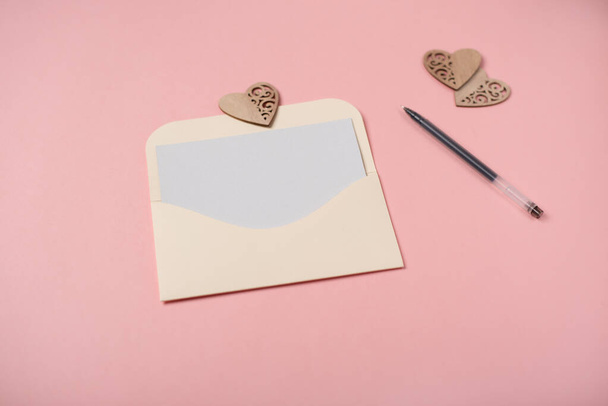 Craft envelope with a blank sheet of paper inside and red wooden heart on the rose background. Romantic love letter for the Valentine's day concept. Space for text. - Фото, зображення