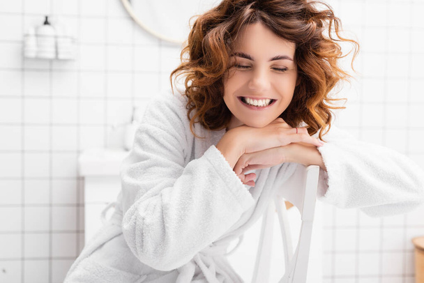 Frau im Bademantel lächelt mit geschlossenen Augen auf Stuhl im Badezimmer  - Foto, Bild