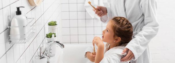 Niño en albornoz sosteniendo el cepillo de dientes cerca de la madre y fregadero en el baño, pancarta  - Foto, Imagen