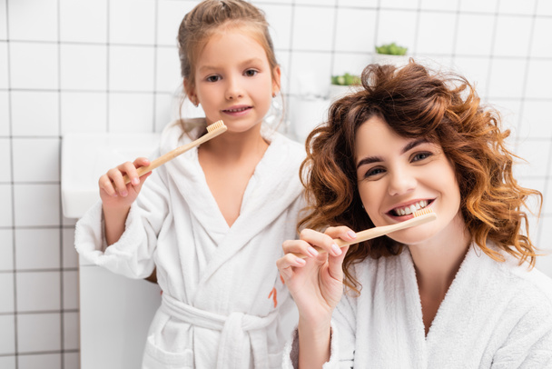 Sorridente donna lavarsi i denti vicino figlia su sfondo sfocato in bagno  - Foto, immagini