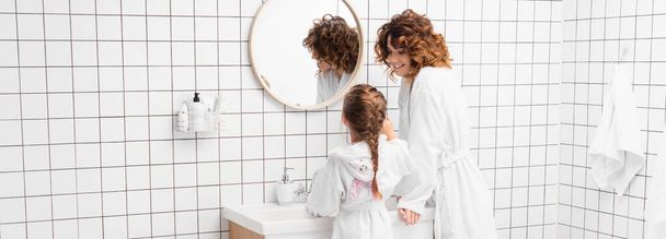 Smiling woman in bathrobe looking at daughter near sink in bathroom, banner  - Foto, immagini