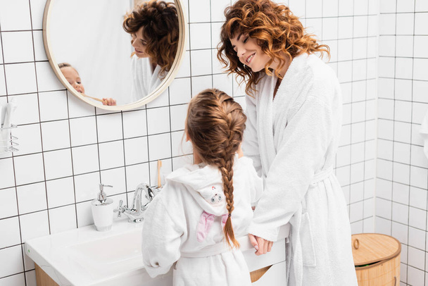 Sorridente madre guardando bambino lavarsi i denti vicino lavandino e specchio  - Foto, immagini