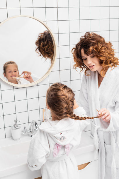 Woman touching hair of daughter brushing teeth near sink in bathroom  - Fotoğraf, Görsel