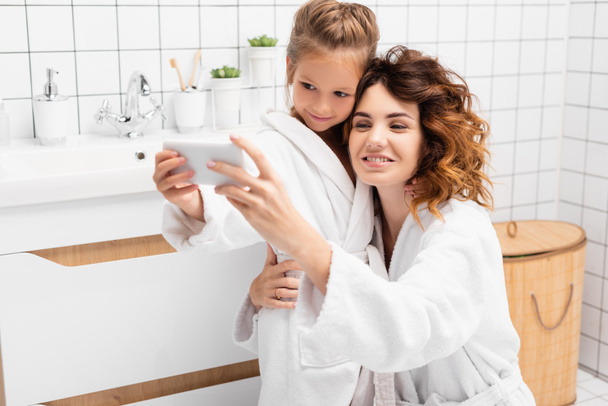 Smiling child taking selfie on smartphone with mother in bathroom  - Fotoğraf, Görsel