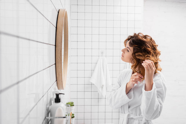 Curly woman in bathrobe adjusting hair near mirror in bathroom  - Foto, Bild