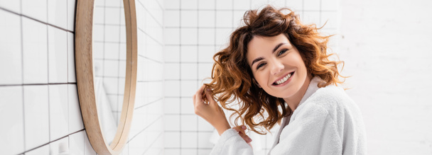 Mulher alegre em roupão de banho branco ajustando o cabelo perto do espelho no banheiro, banner  - Foto, Imagem