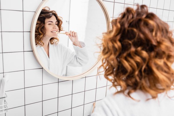 Woman in bathrobe brushing teeth near mirror on blurred foreground  - Fotografie, Obrázek