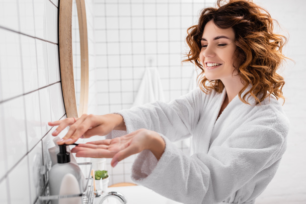 Cheerful woman applying cosmetic cream on blurred foreground in bathroom  - Fotó, kép