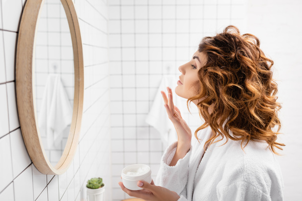 Side view of curly woman applying face cream near mirror in bathroom  - Photo, Image