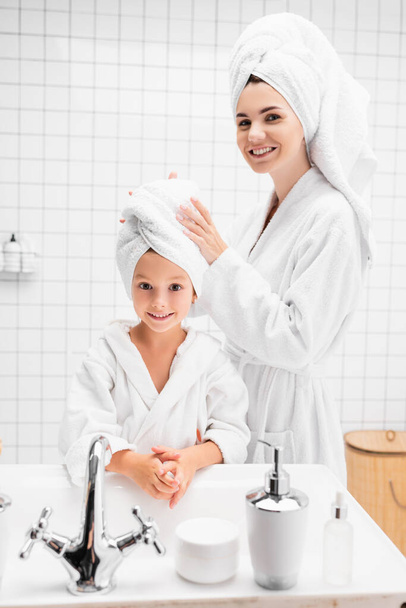Madre sorridente in accappatoio che indossa un asciugamano sulla testa del bambino in bagno  - Foto, immagini