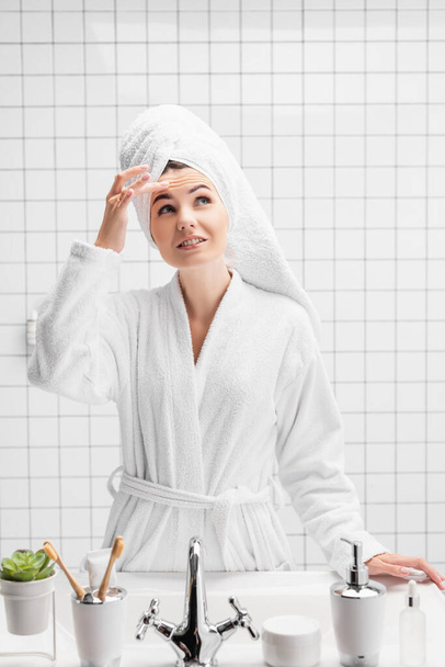 Улыбающаяся женщина в полотенце касается лба в ванной комнате  - Фото, изображение