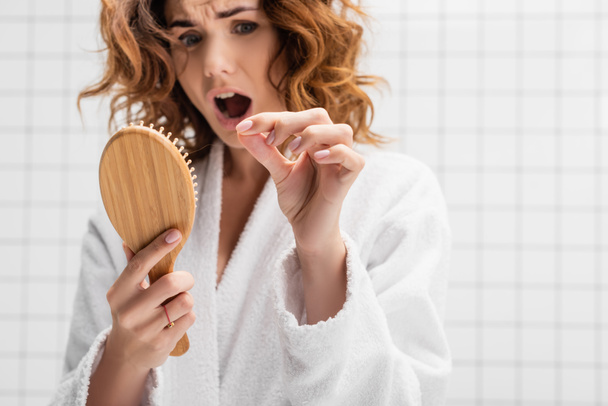 Cepillo de pelo en la mano de la mujer sorprendida sobre fondo borroso en el baño sobre fondo borroso - Foto, Imagen