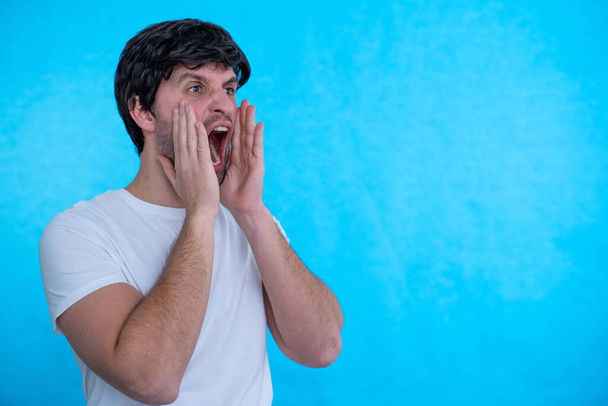 Man standing over blue background Shouting angry out loud with hands over mouth - Fotó, kép