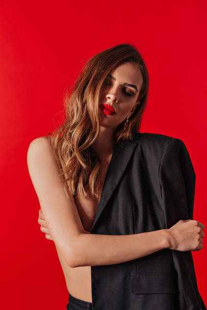 Stylish charming lady with red lips and wavy hairs covering black jacket posing over red background. Valentine's day. High quality photo - Φωτογραφία, εικόνα