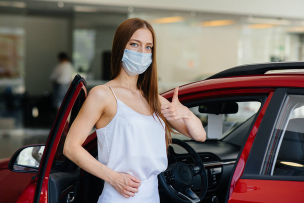 A young pretty girl inspects a new car at a car dealership in a mask during the pandemic. The sale and purchase of cars, in the period of pandemia - Foto, Imagem